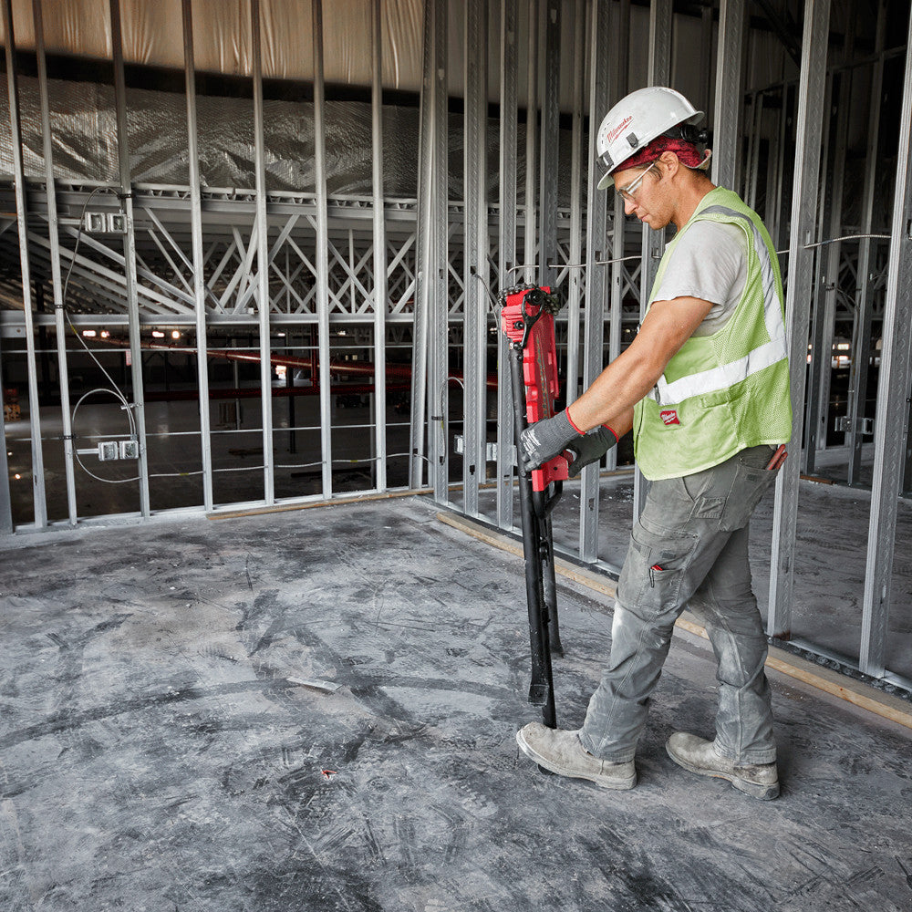 Worker in safety gear holding red Milwaukee chain vise with tripod stand on construction site floor.