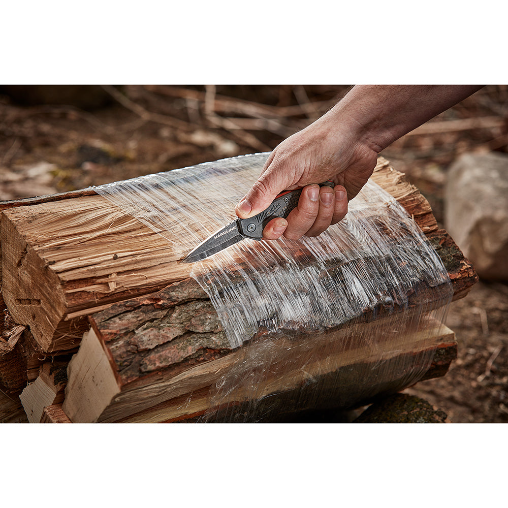 Person holding open pocket knife with black handle and blade against wooden log.