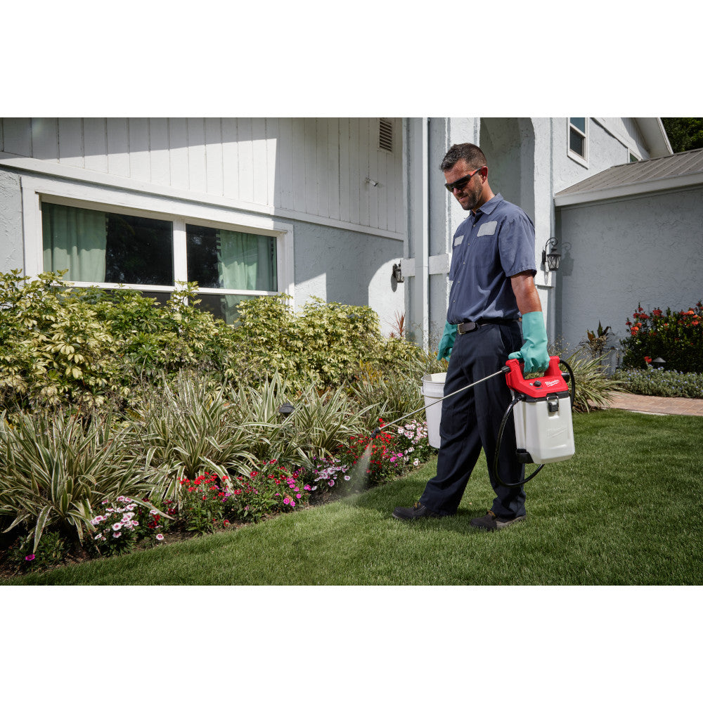White handheld sprayer tank with red pump handle being held by person in yard near landscaping.