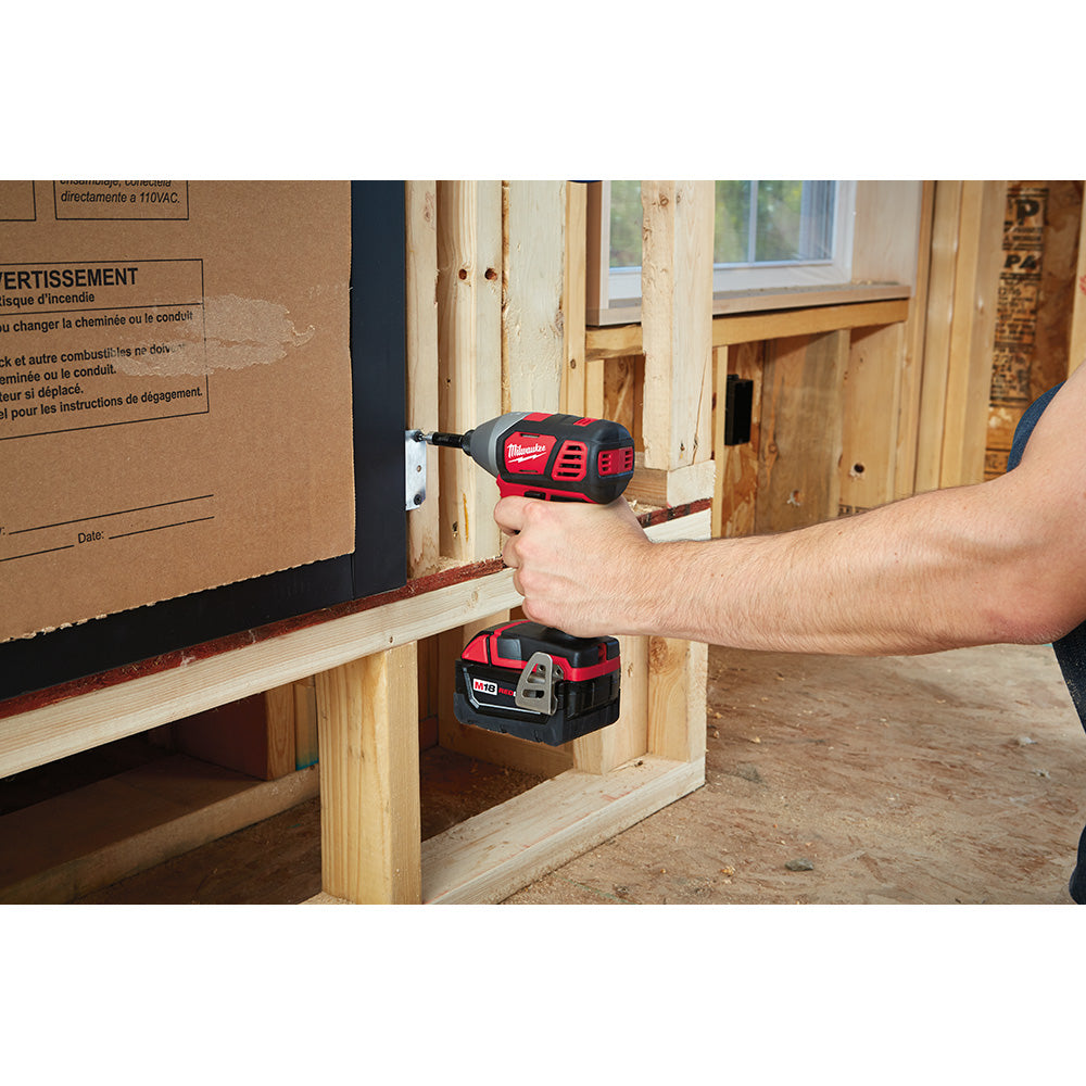 Red and black Milwaukee M18 hex impact driver held in hand, driving fastener into wooden framing on construction site.