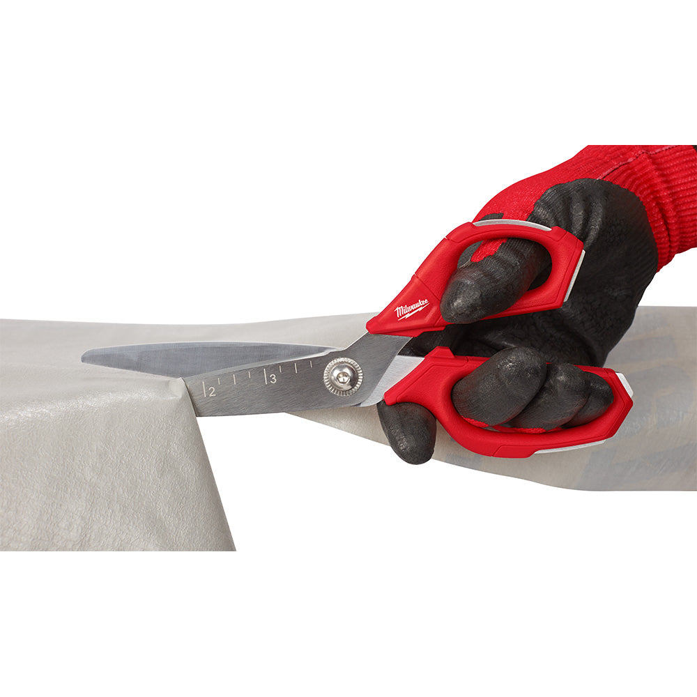 Red and black straight scissors with metal blades and measurement markings, held in a gloved hand cutting through material.