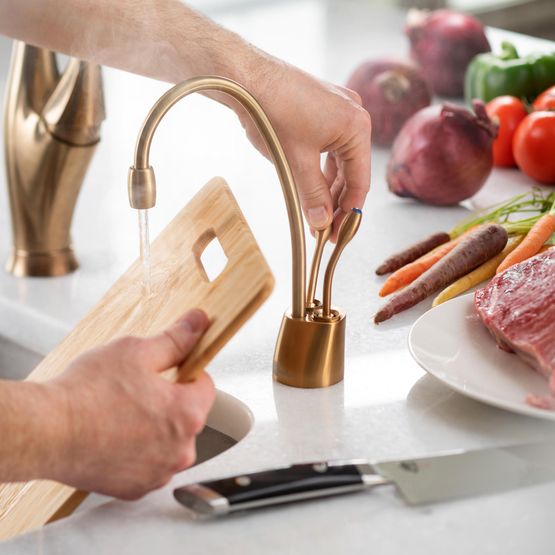 Brushed bronze instant hot and cold faucet with curved spout dispensing water onto wooden cutting board in kitchen.