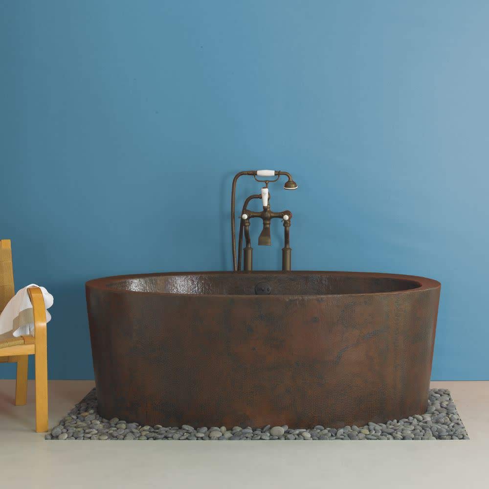 Hammered copper freestanding bathtub with textured surface and matching bronze floor-mounted filler and hand shower fixture.