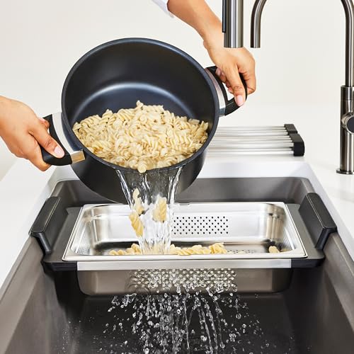 Stainless steel sink colander with perforated surface catching drained pasta from black pot over sink.