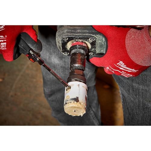 Hole saw bit installed in drill chuck, showing metal cutting head and white arbor sleeve with red gloved hands holding tool.