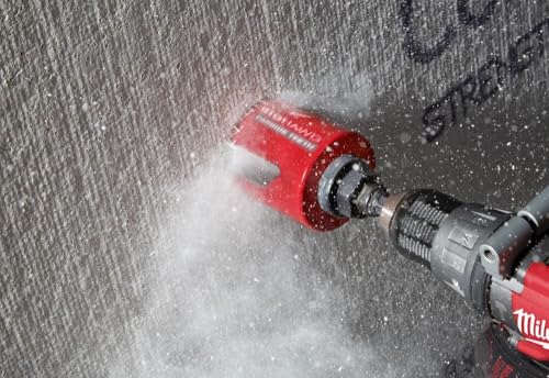 Red Milwaukee hole saw with carbide teeth drilling into concrete surface with water spray.