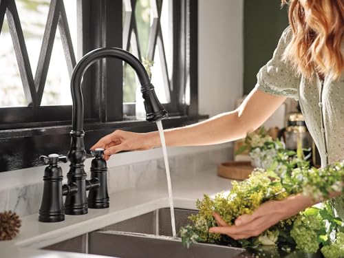 Matte black two-handle bridge kitchen faucet with pulldown sprayer over white sink basin.