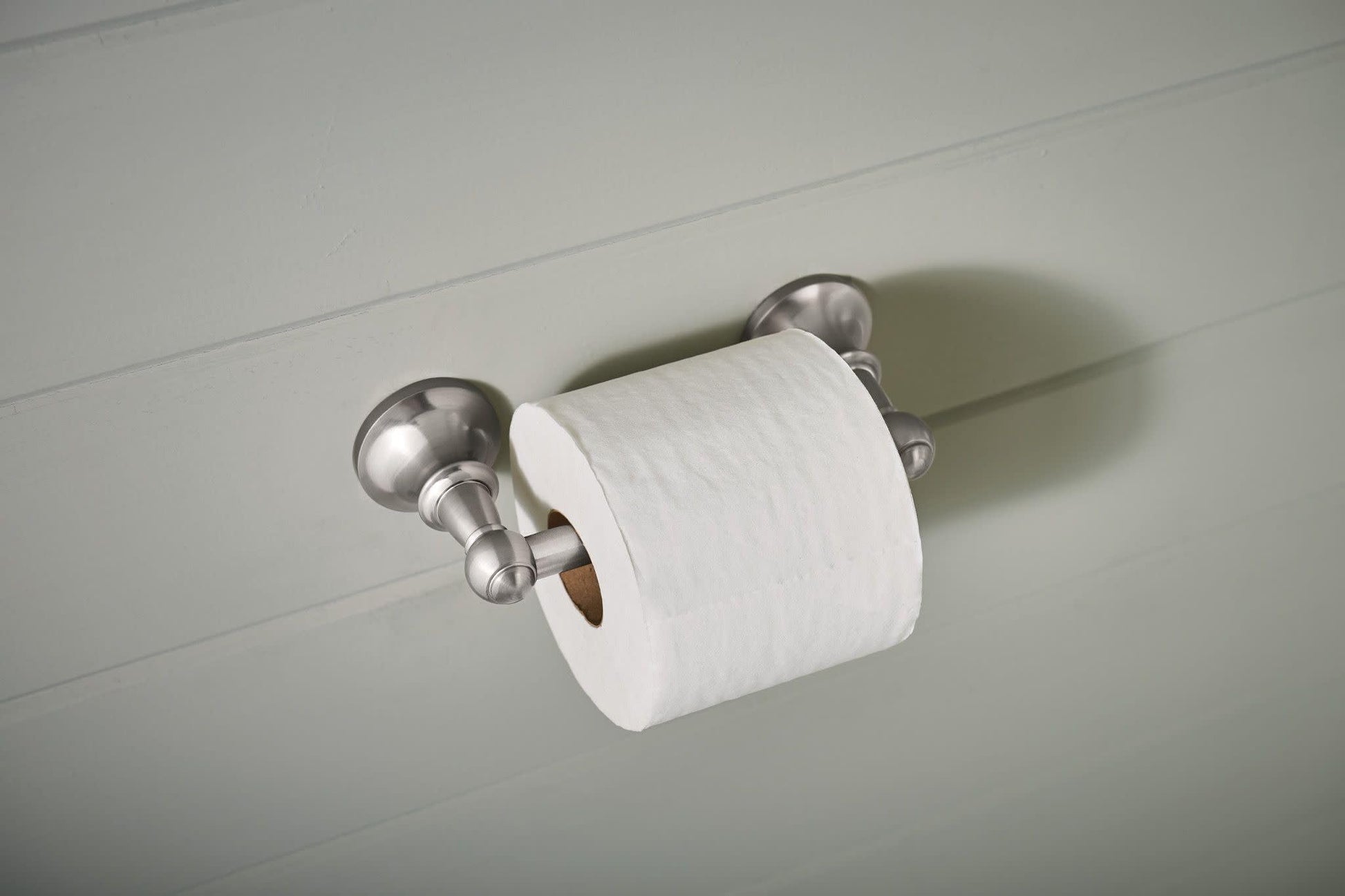 Brushed nickel pivoting toilet paper holder mounted on white wall with white toilet paper roll installed.