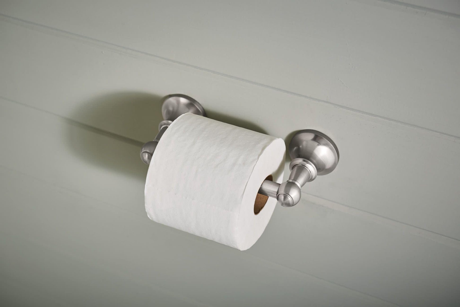 Brushed nickel toilet paper holder with pivoting arms mounted on white wall with white roll installed.