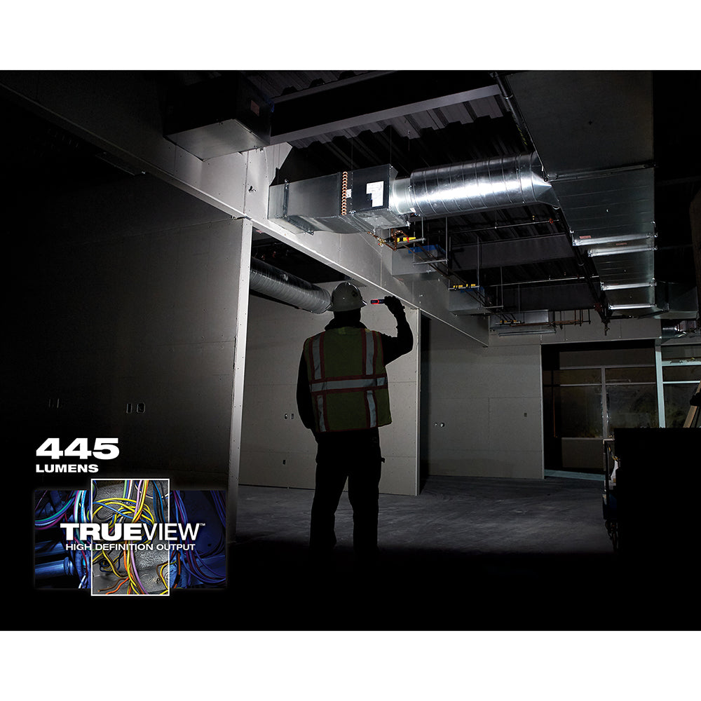 Worker in safety vest holding compact flood light illuminating industrial workspace at night.