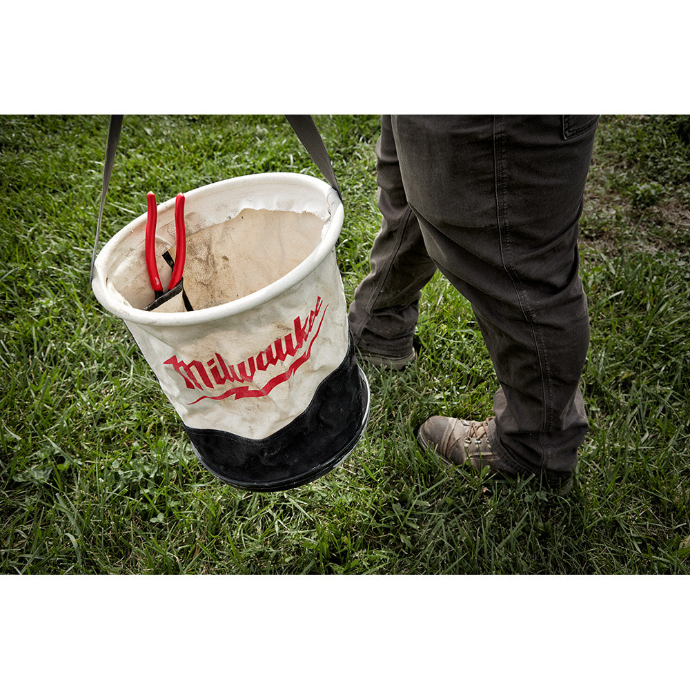 Cream canvas bucket with red Milwaukee branding and red handles, partially filled with sand, held by person in dark pants.