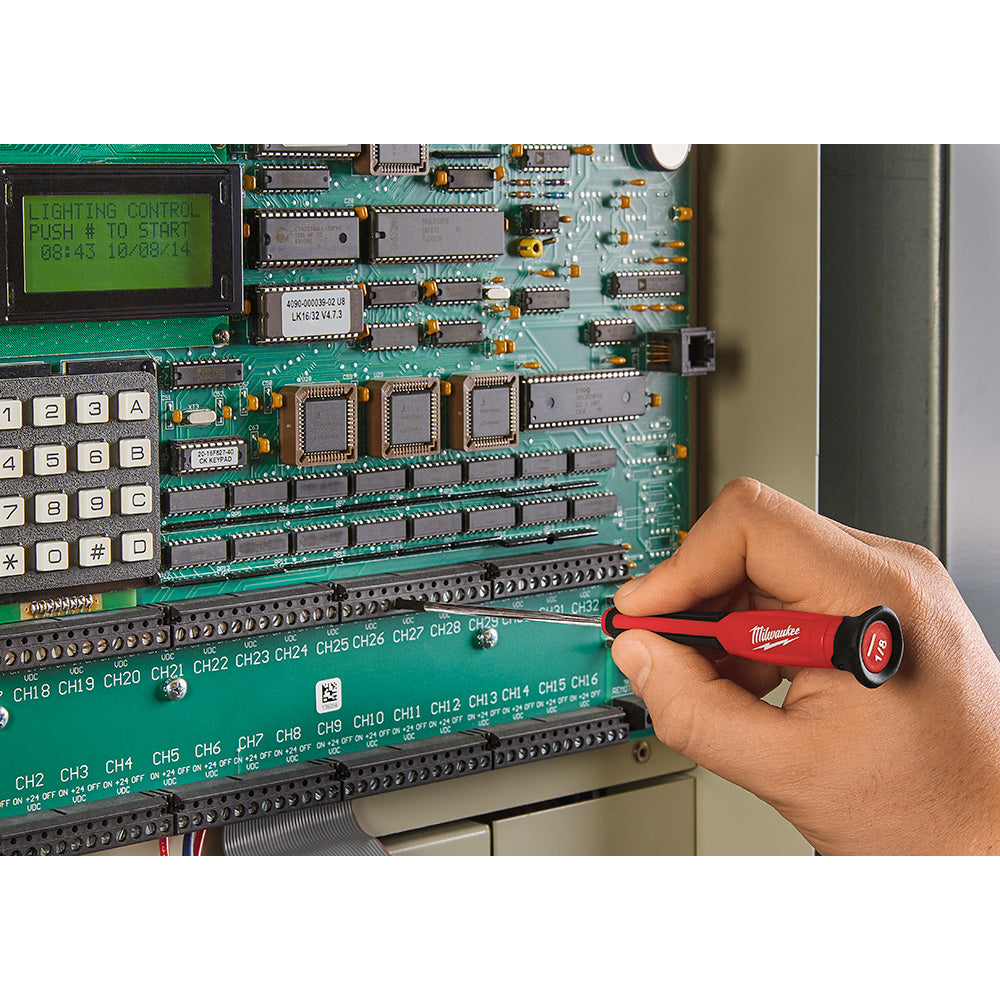 Hand holding red Milwaukee precision screwdriver with black grip working on green circuit board.