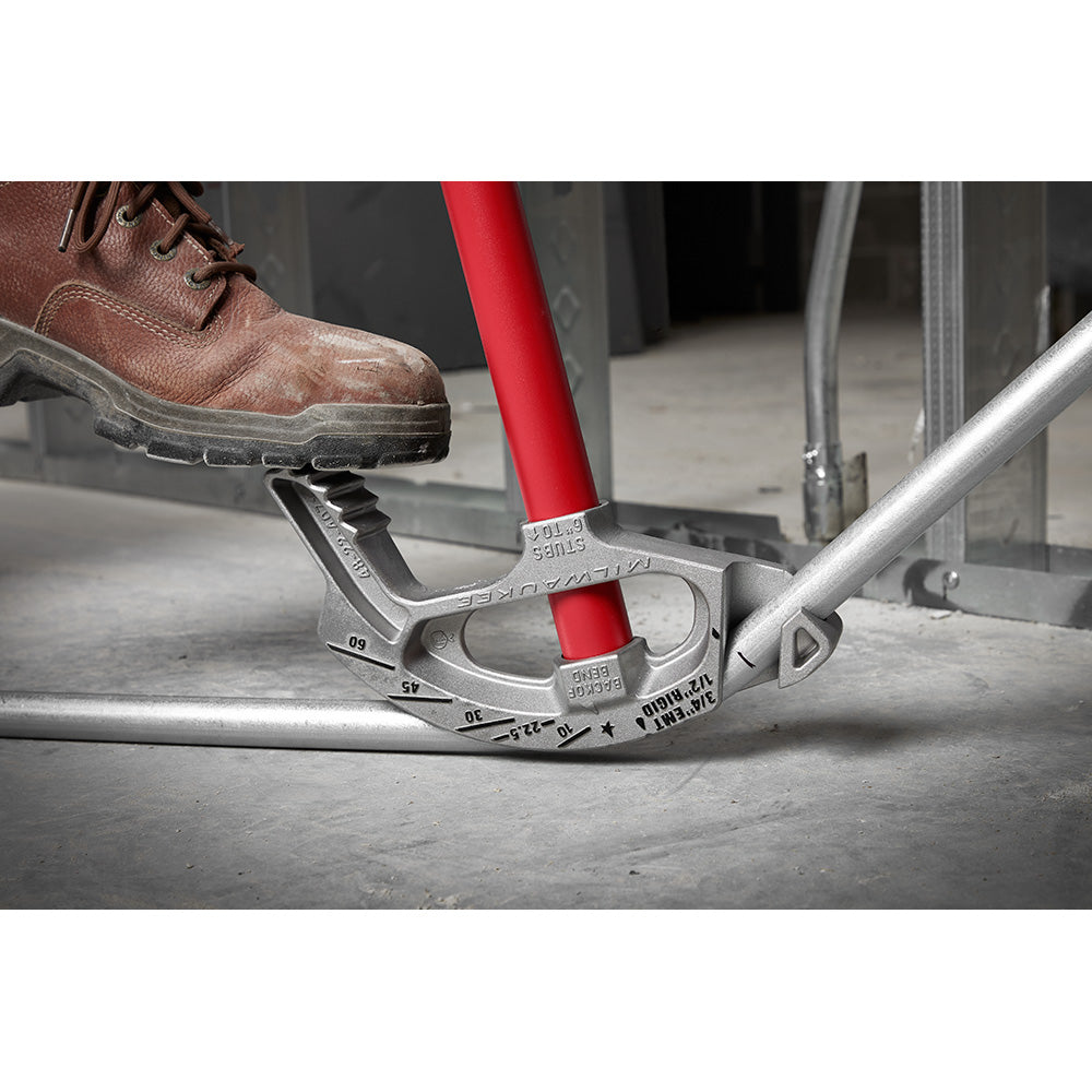 Aluminum conduit bender with red handle and measurement markings, held against metal conduit on concrete surface with worker'