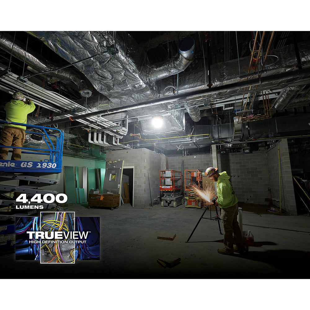 Milwaukee M18 RADIUS LED compact site light mounted on tripod illuminating an indoor construction area with workers.