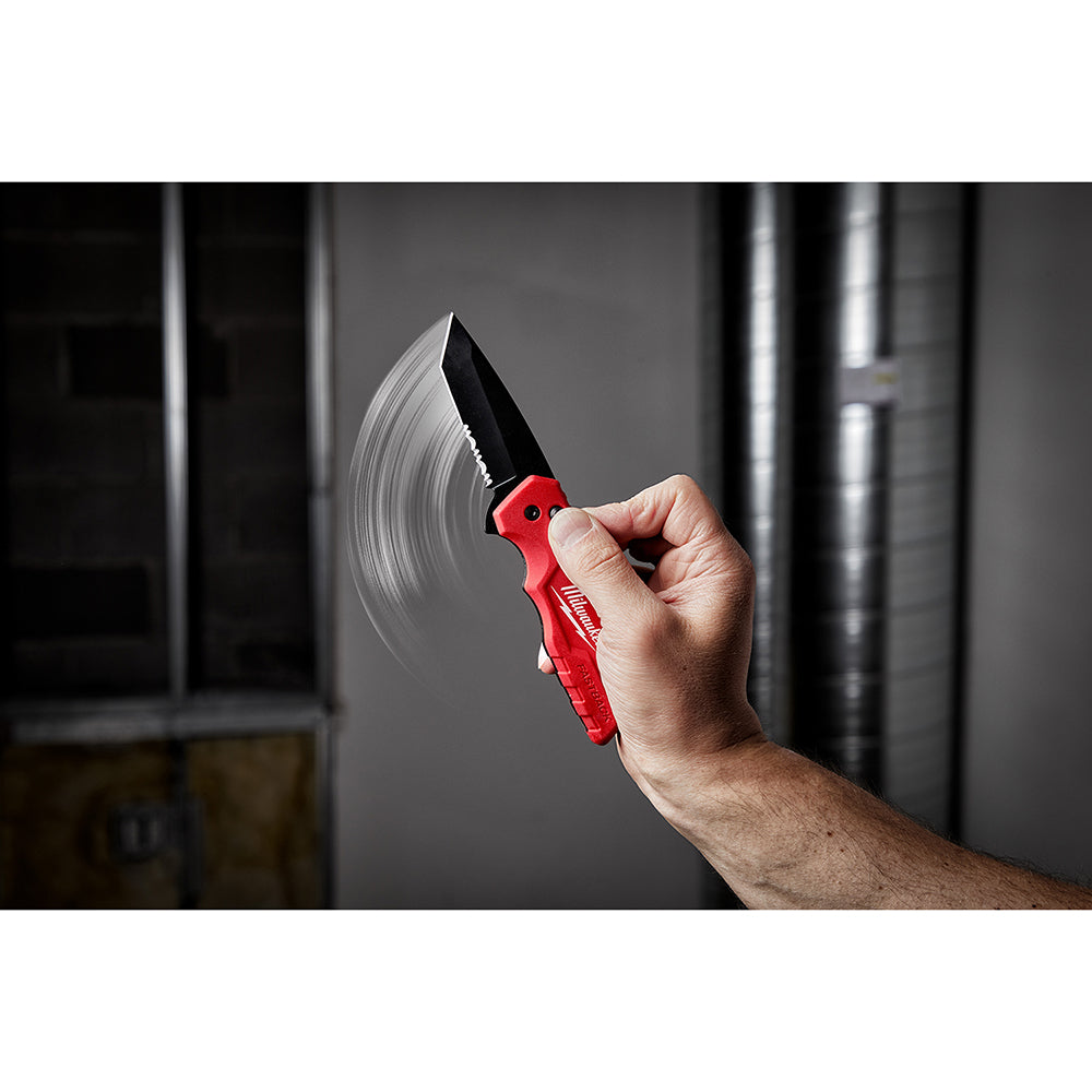 Red and black folding knife with serrated blade held open in hand against industrial background.