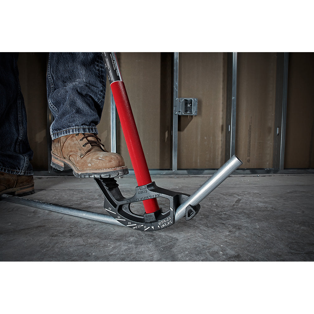 Red and black metal conduit bender with long handle being used to bend metallic pipe on concrete floor.