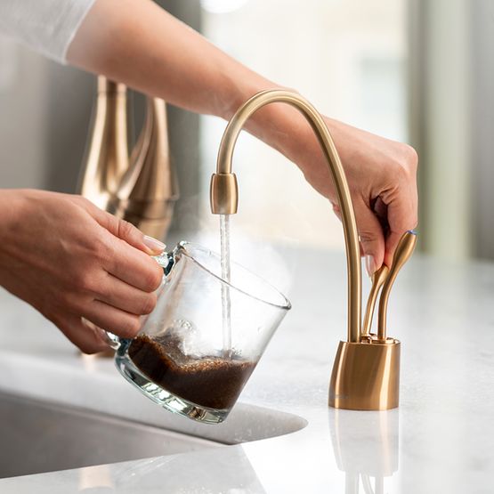 Brushed bronze instant hot and cold faucet with curved spout dispensing dark liquid into a glass held by a hand.
