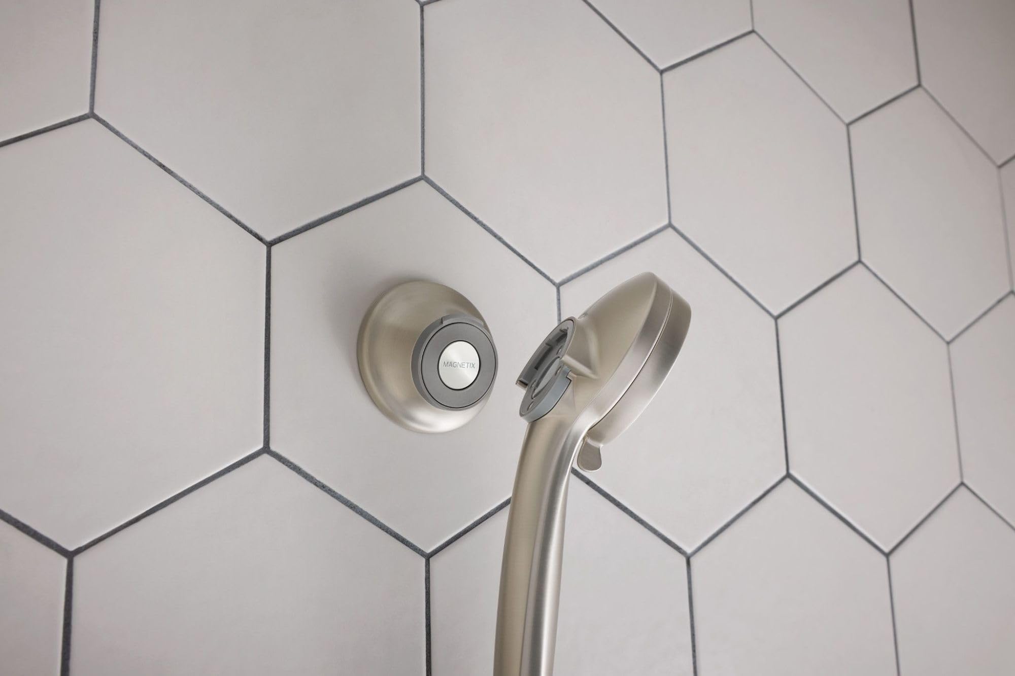 Brushed nickel showerhead with magnetic remote cradle mounted on hexagonal tile wall.