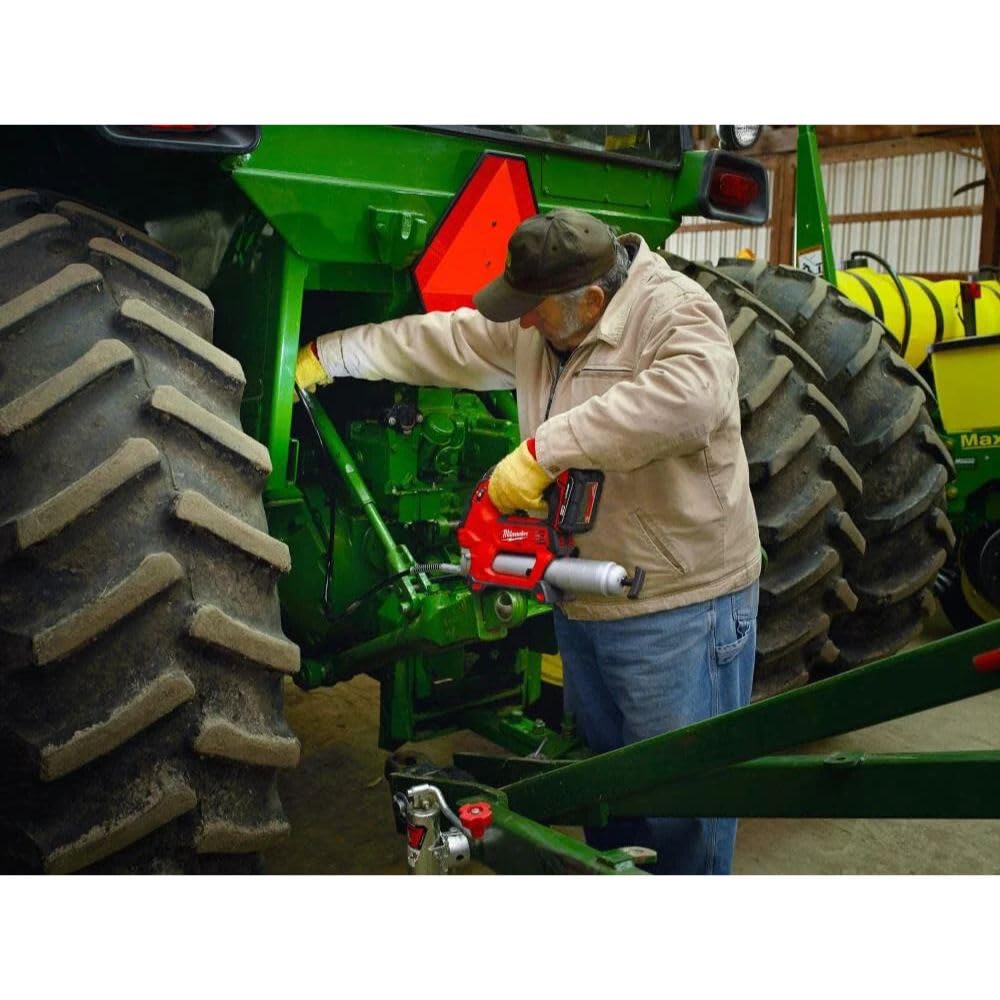 Red and black Milwaukee M18 cordless grease gun being used on farm equipment by a worker in a barn.