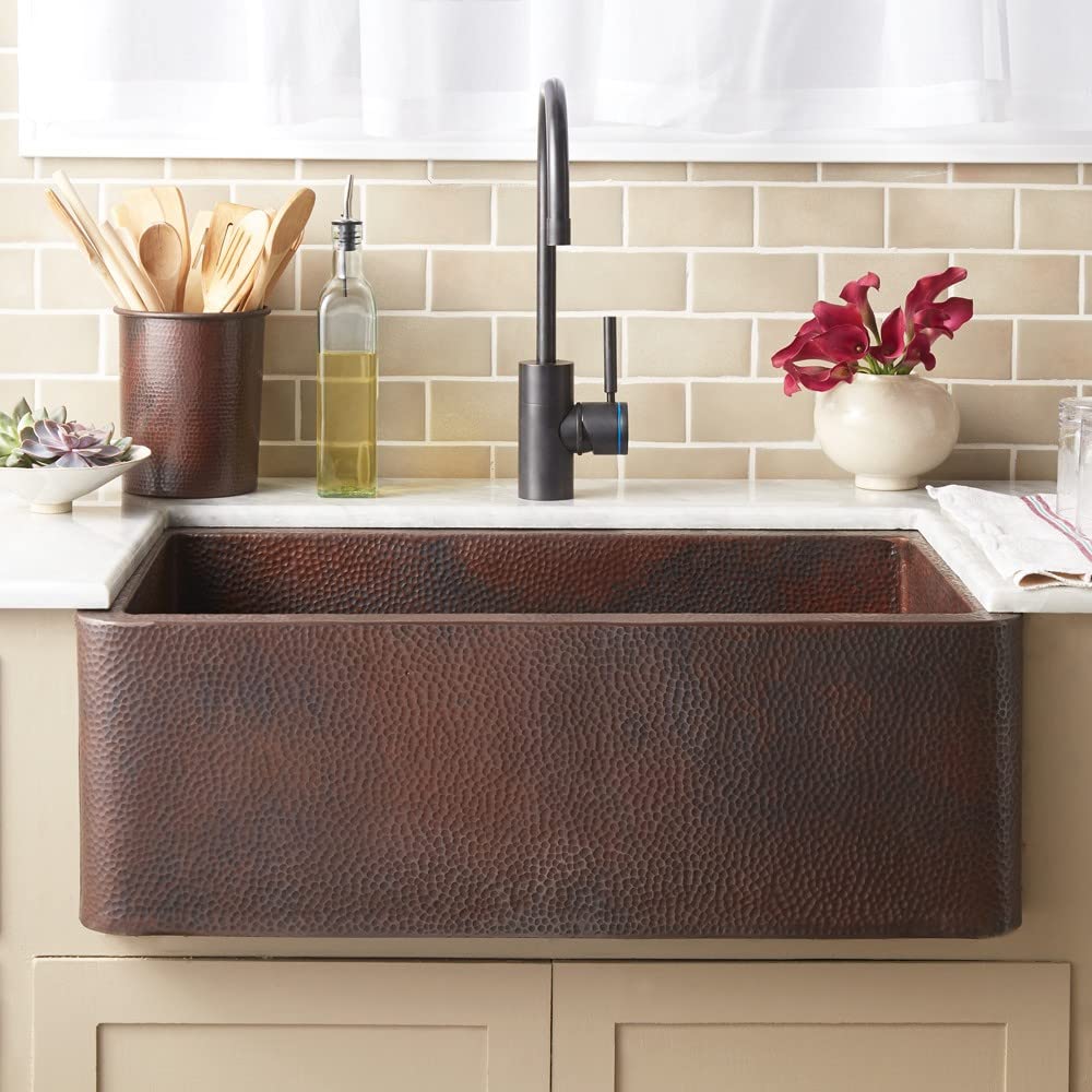 Antique copper farmhouse sink with hammered texture, mounted on beige cabinet below subway tile backsplash, paired with black