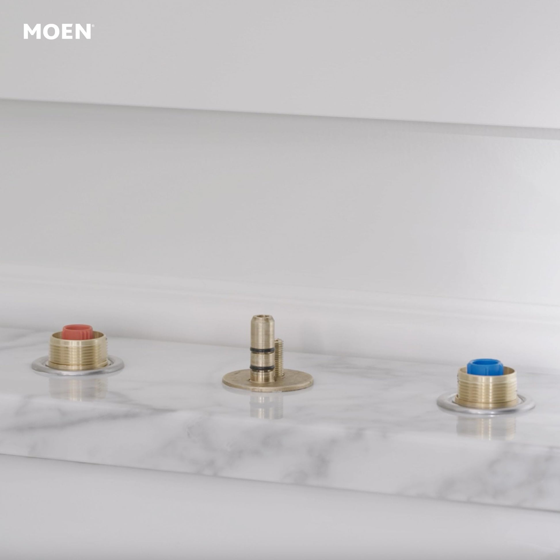 Moen rough-in valve assembly with three brass components featuring red and blue valve caps on marble surface.