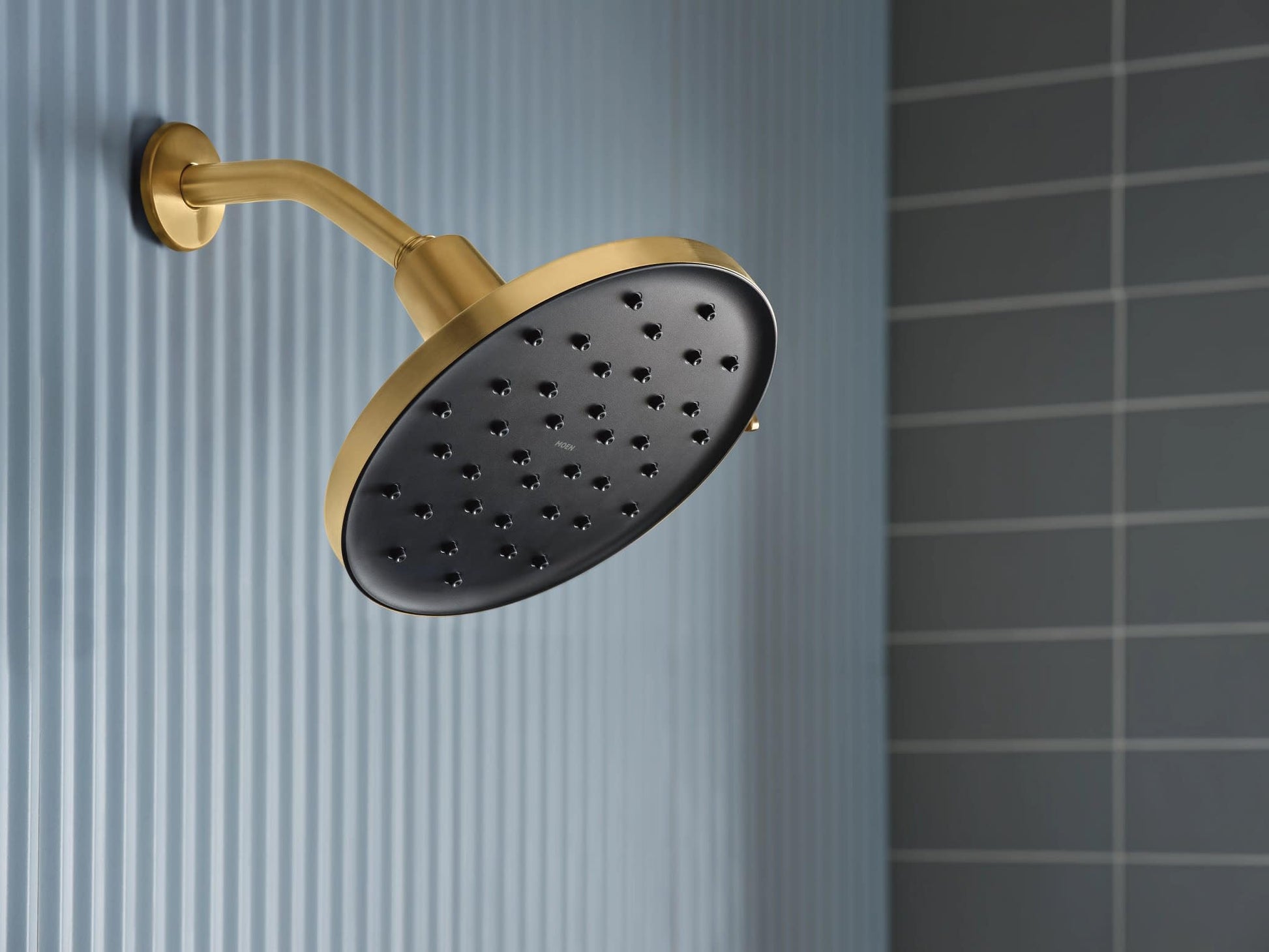 Brushed gold rainshower head with black spray face and curved arm mounted on wall.