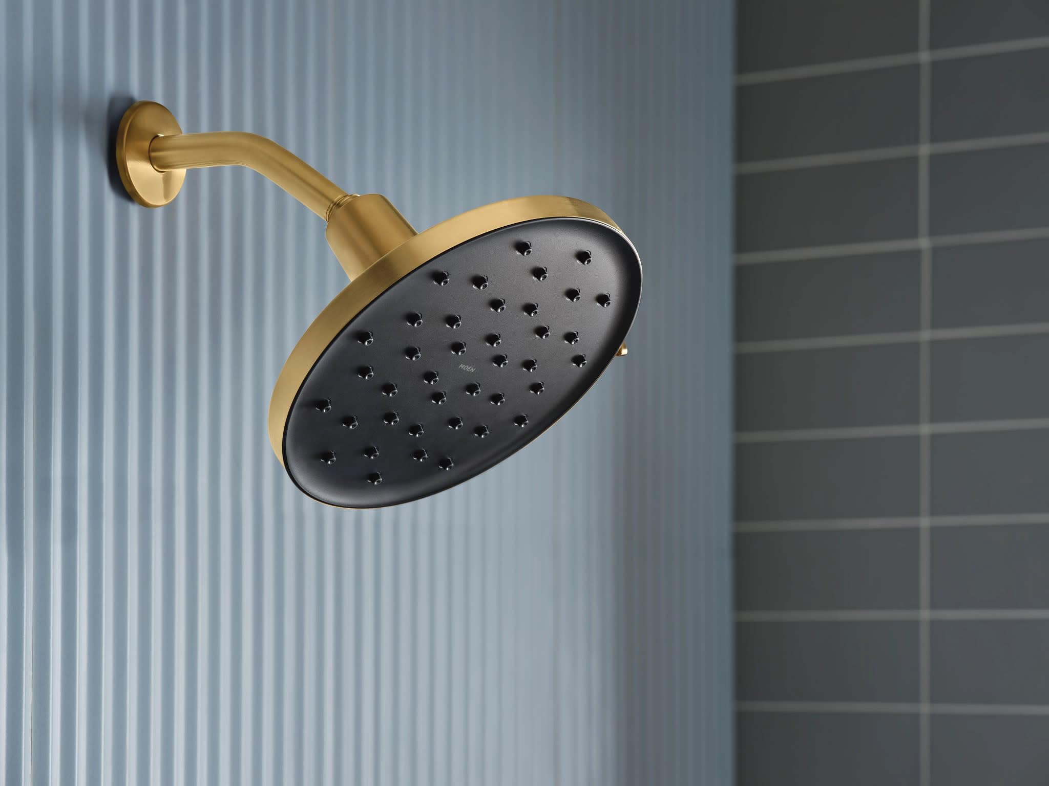 Brushed gold rainshower head with black spray face and curved arm mounted on wall.