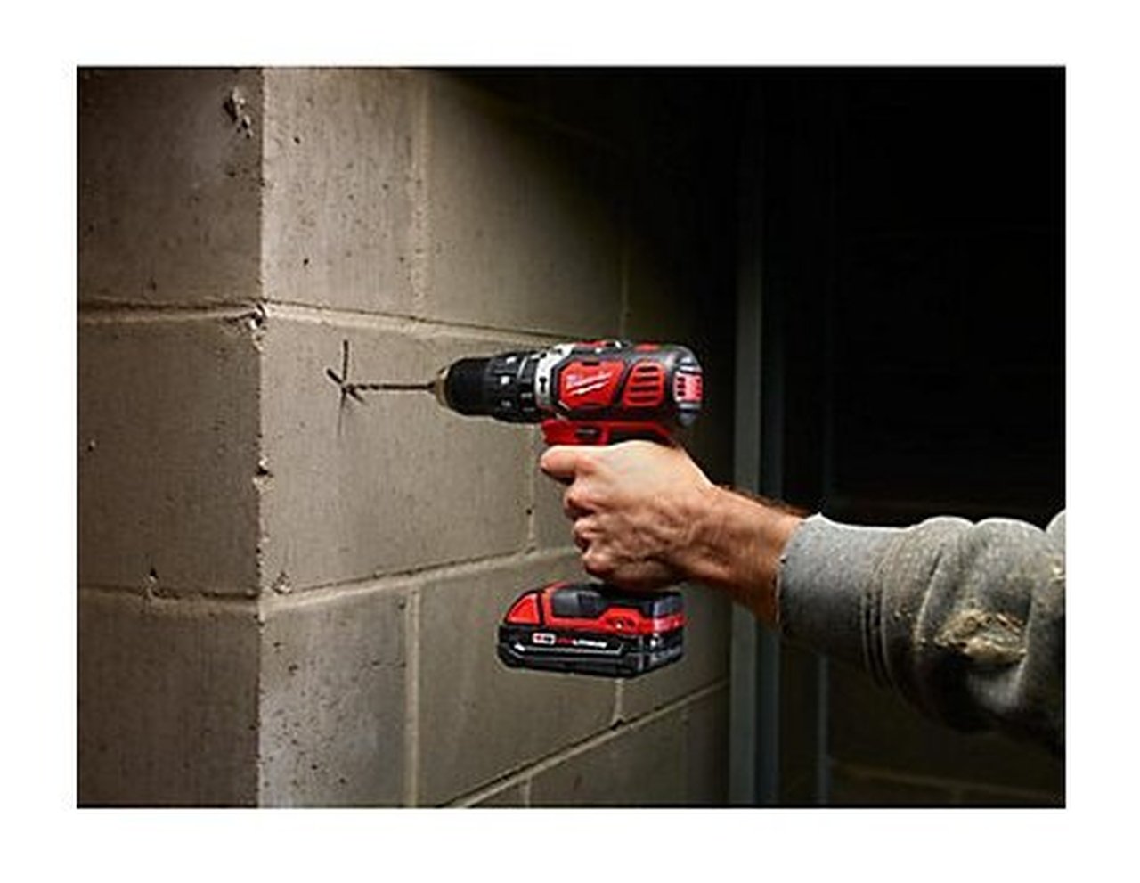 Red and black cordless drill being held against concrete wall with battery pack visible.