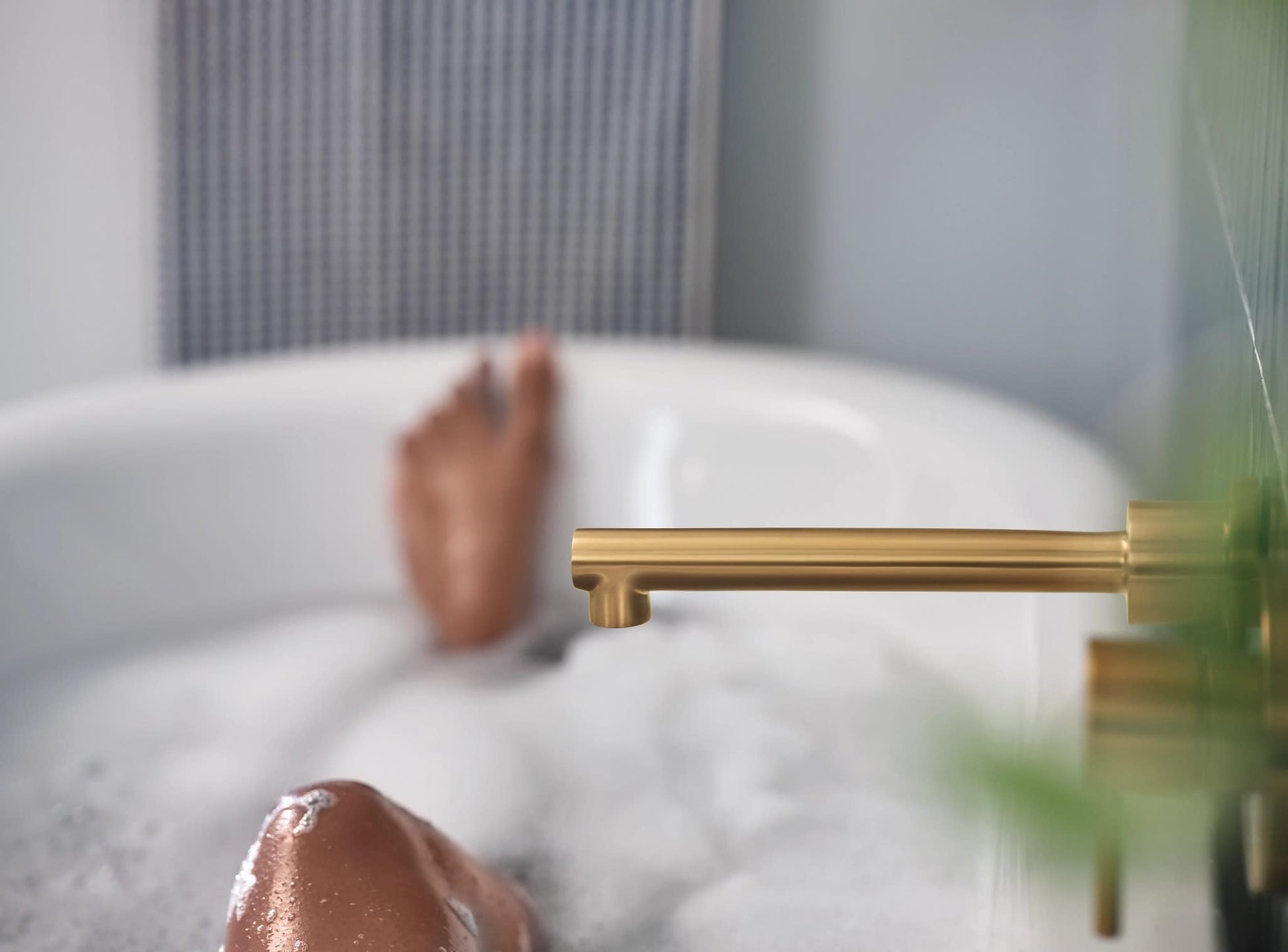Brushed gold wall-mounted tub faucet with single handle and extended spout over a white bathtub.