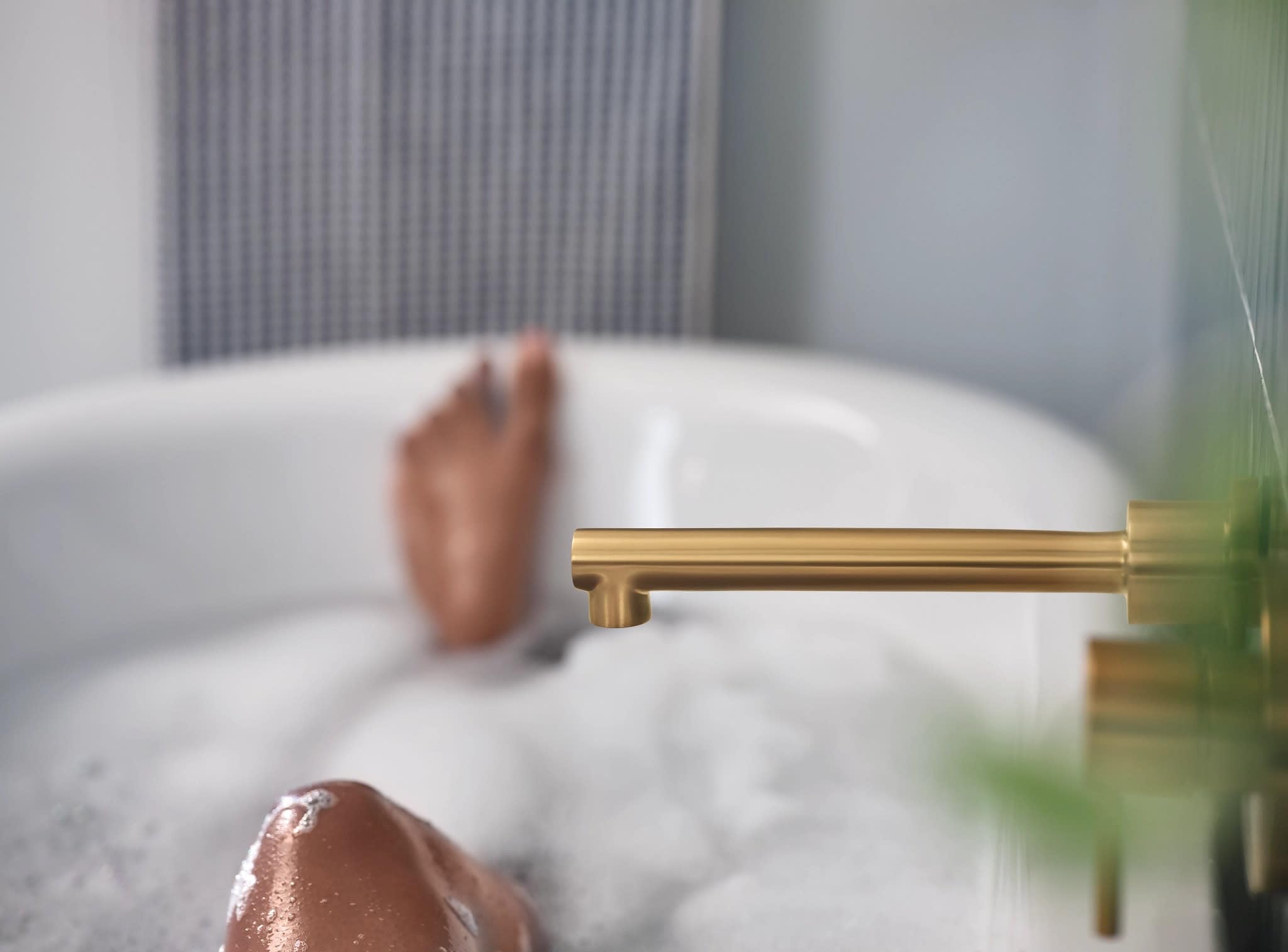 Brushed gold wall-mounted tub faucet with single handle and extended spout over a white bathtub.