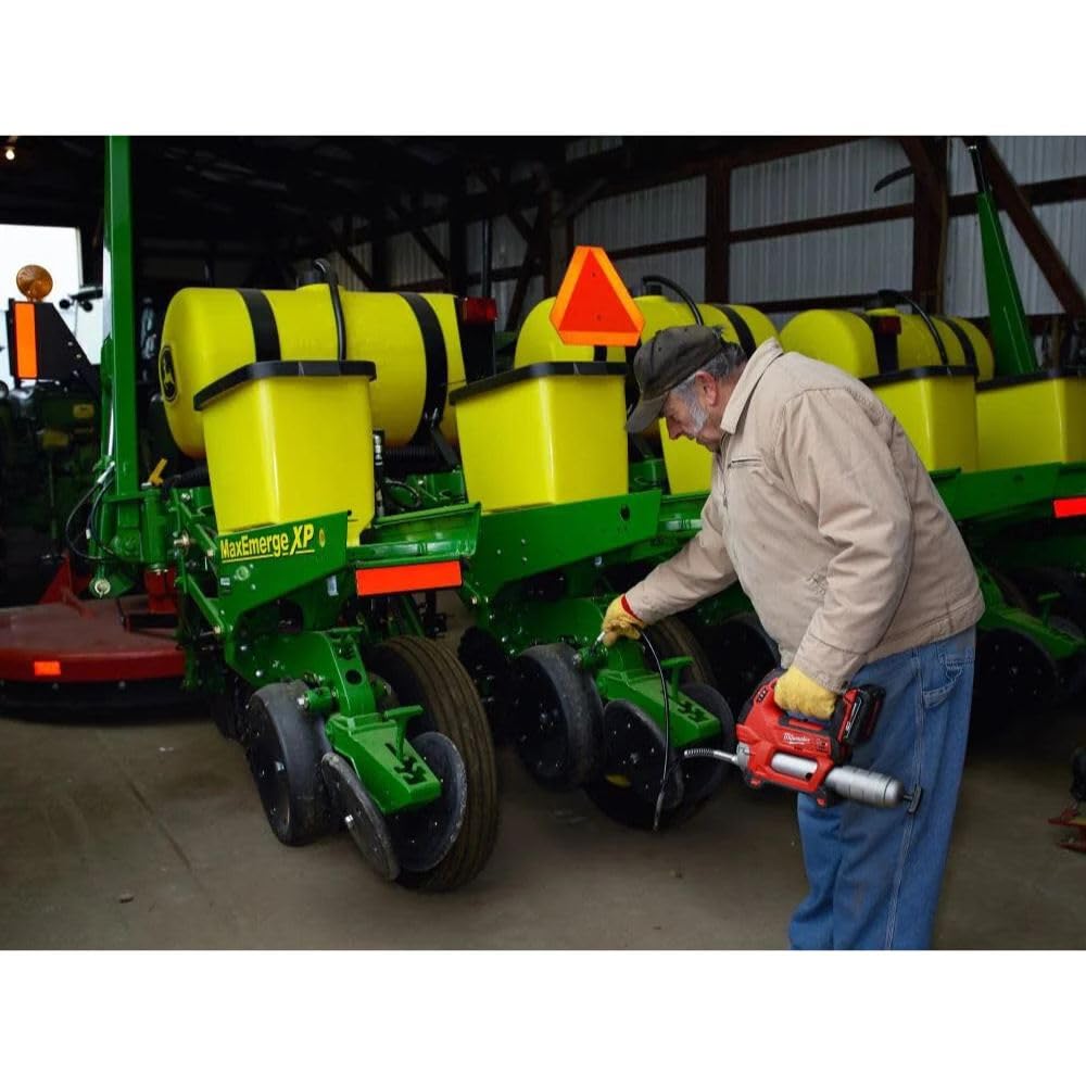 Worker in beige jacket using red and black Milwaukee grease gun on green agricultural equipment in warehouse.