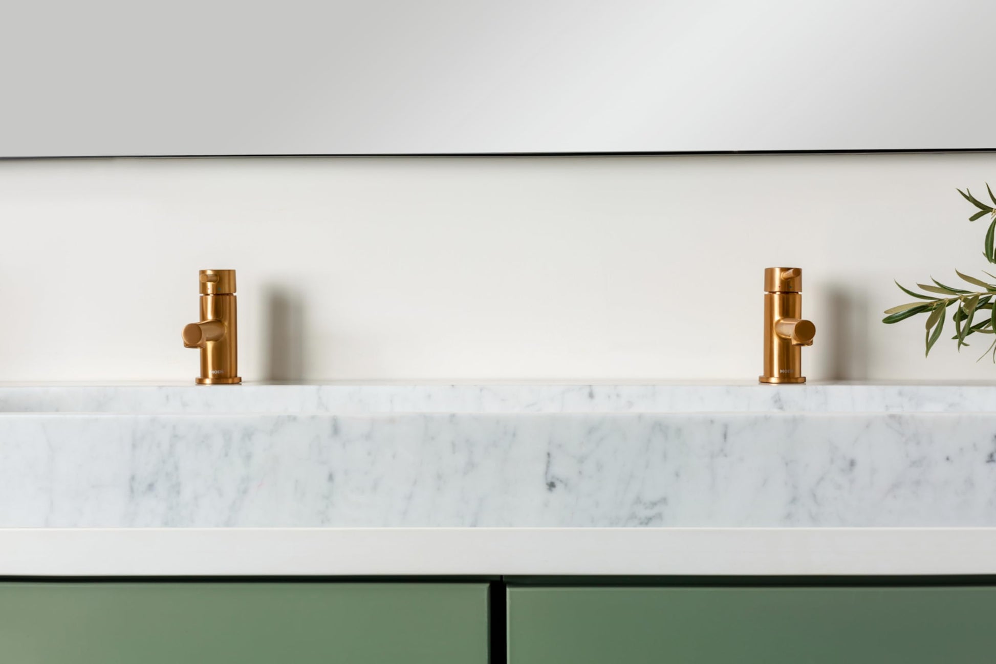 Two brushed gold single-handle low-arc faucets mounted on white marble countertop with green cabinetry below.
