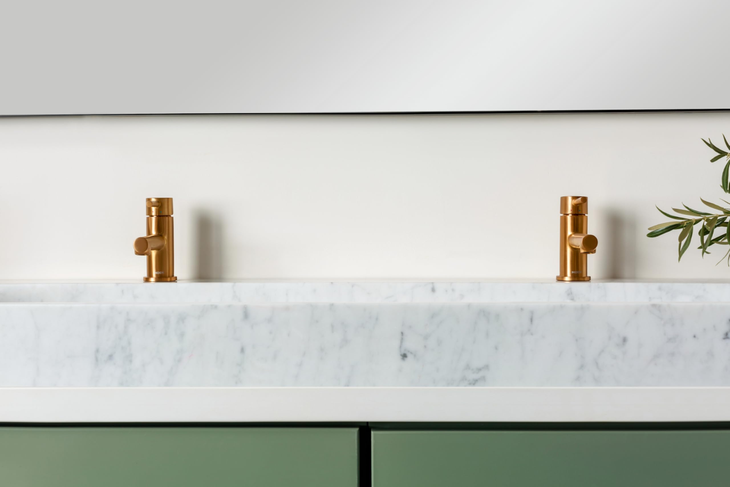Two brushed gold single-handle low-arc faucets mounted on white marble countertop with green cabinetry below.