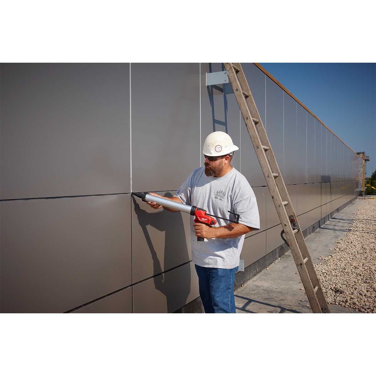 Worker using red and black cordless caulk gun to apply sealant to large gray metal panels outdoors.