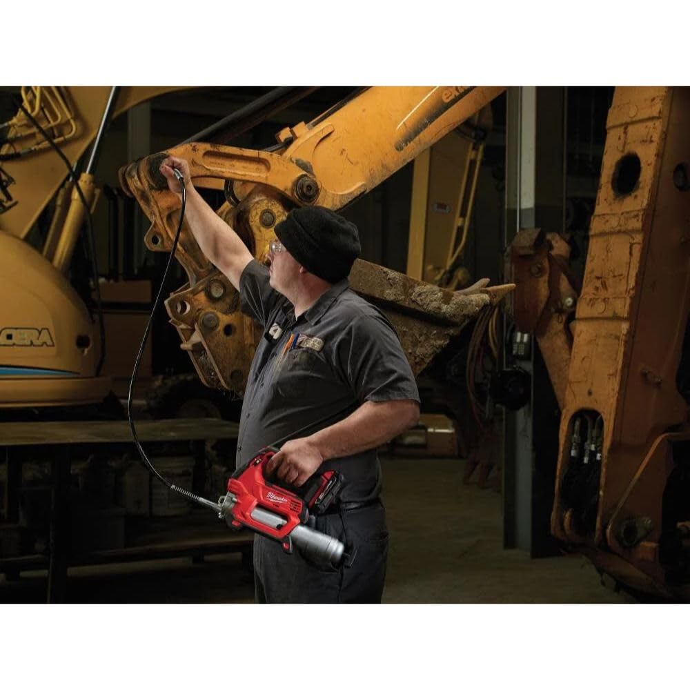 Worker holding red and silver Milwaukee cordless grease gun near heavy machinery in industrial setting.