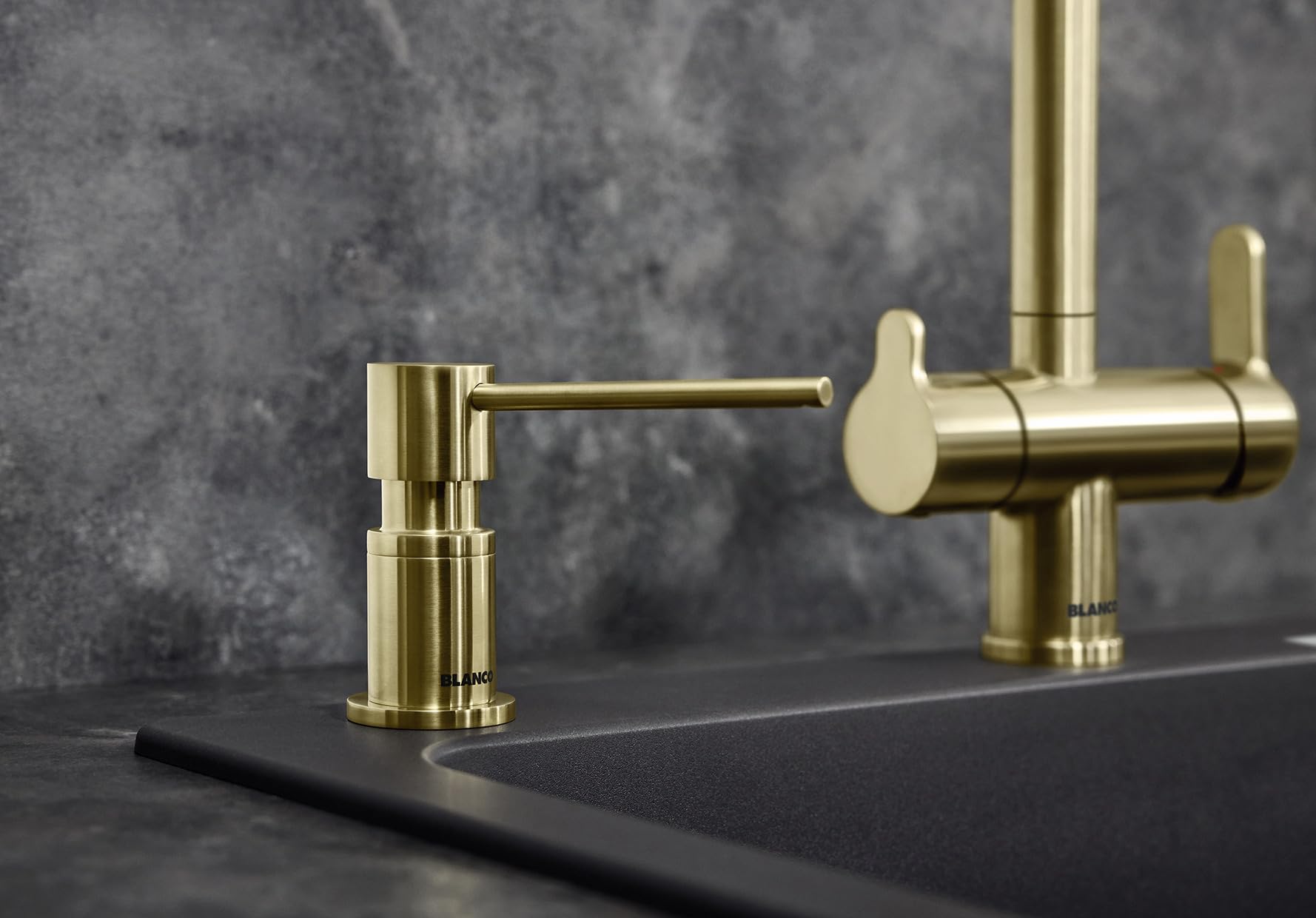 Brass soap dispenser with cylindrical pump and angled nozzle on dark countertop.