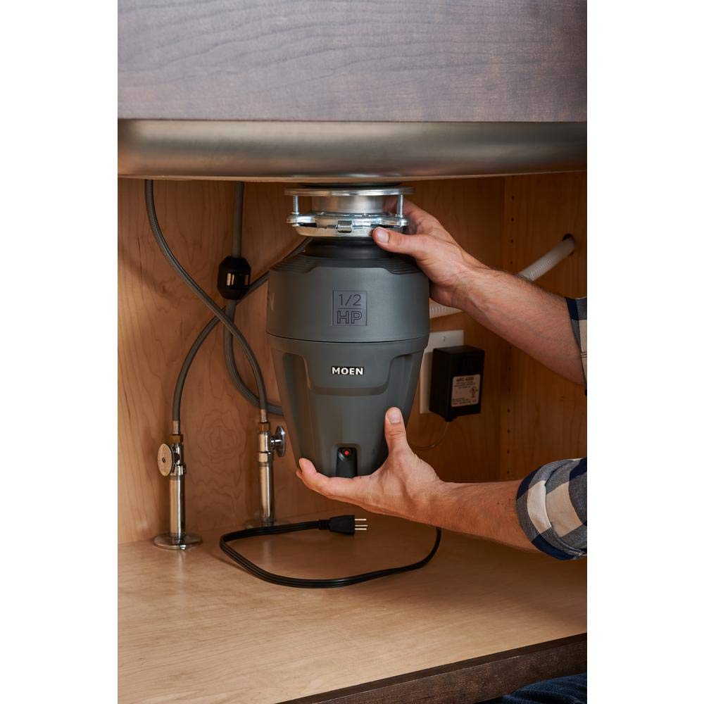 Black Moen 1/2 HP garbage disposal unit mounted under sink with power cord and drain connections visible.