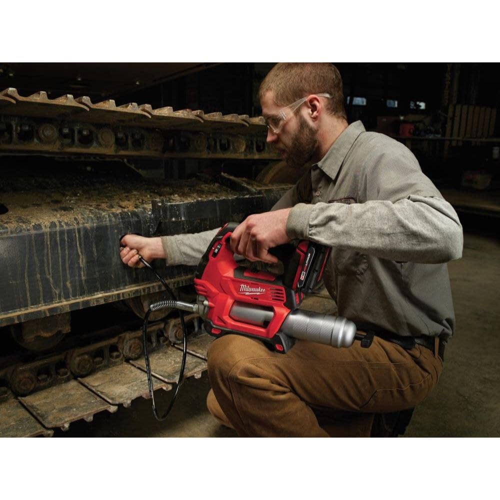 Milwaukee M18 red cordless grease gun with hose attachment being used on machinery by a worker.