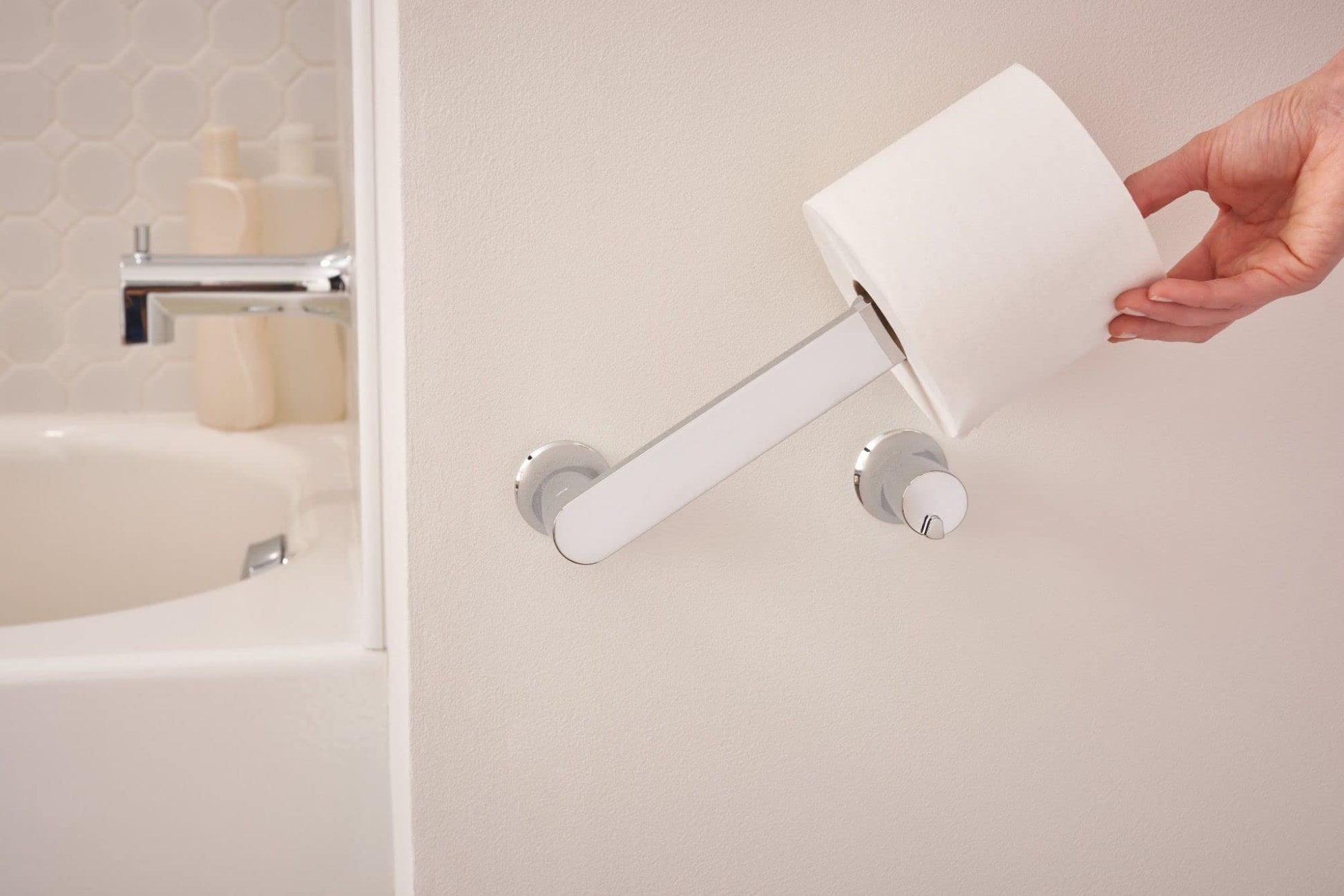 Chrome pivoting toilet paper holder mounted on bathroom wall with white roll loaded and hand pulling it outward.