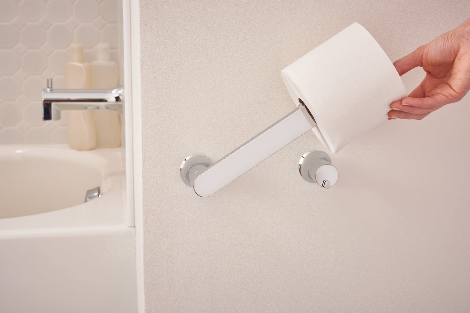 Chrome pivoting toilet paper holder mounted on bathroom wall with white roll loaded and hand pulling it outward.