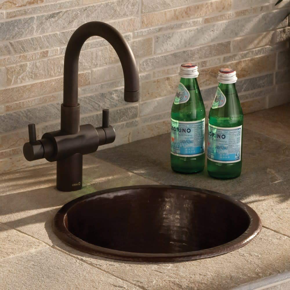 Hammered copper round bar sink with dark bronze faucet and dual handles installed in stone countertop.