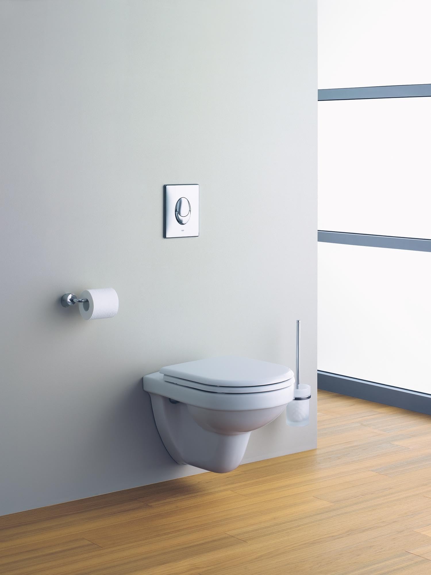 Chrome rectangular wall plate with circular flush button mounted above wall-mounted toilet.