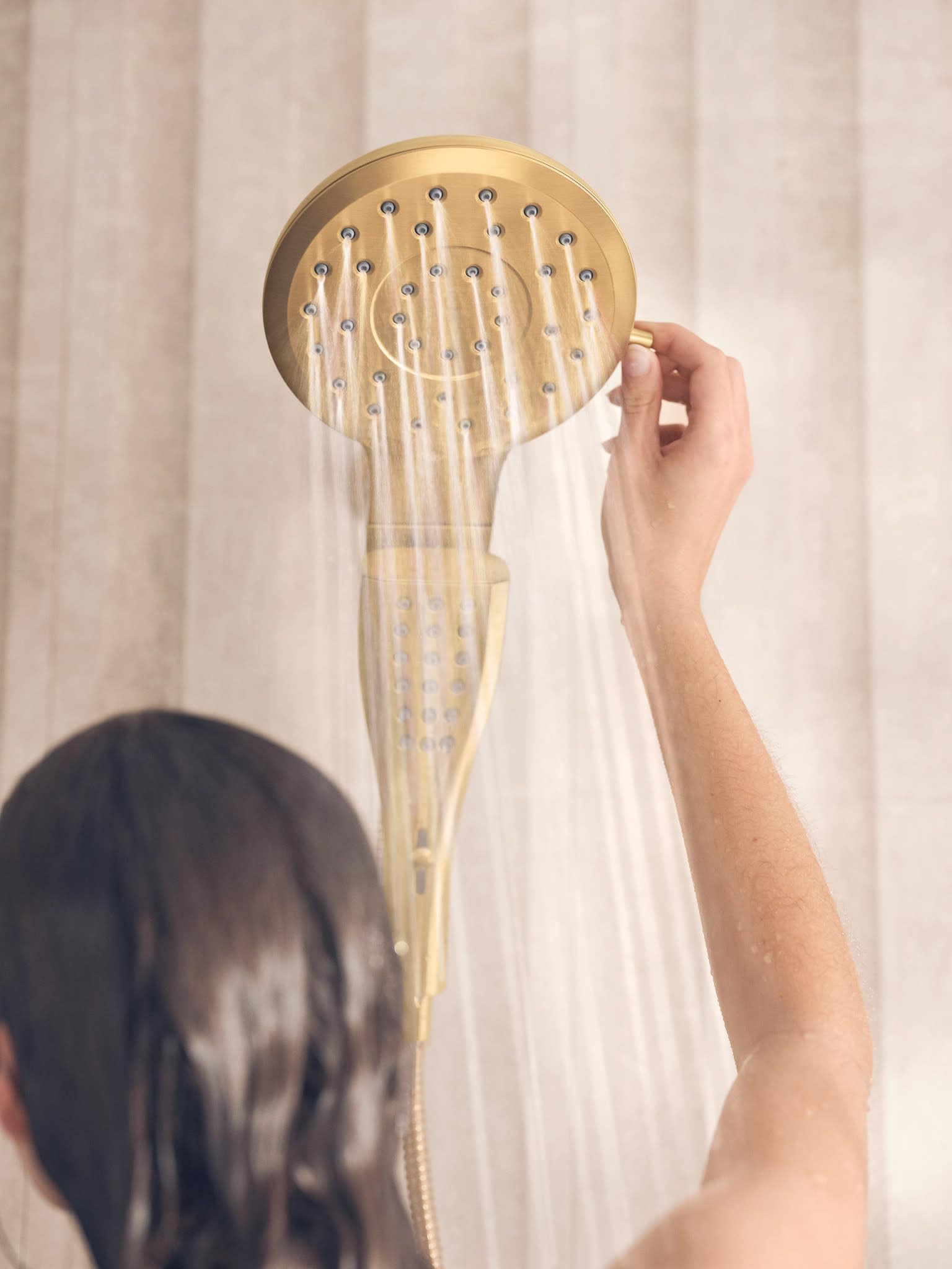 Brushed gold circular showerhead with multiple spray jets, held by a hand demonstrating water flow.