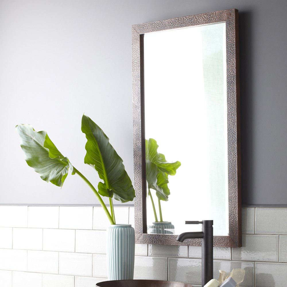 Rectangular mirror with textured antique frame mounted above white subway tile backsplash, reflecting green plant stems and b