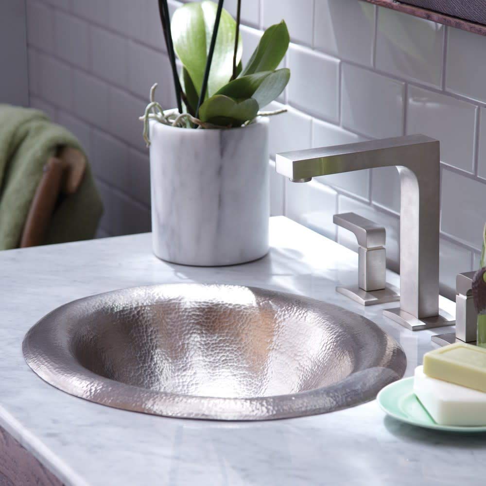 Hammered metal round bathroom sink with lotus-shaped rim and brushed nickel faucet on marble countertop.