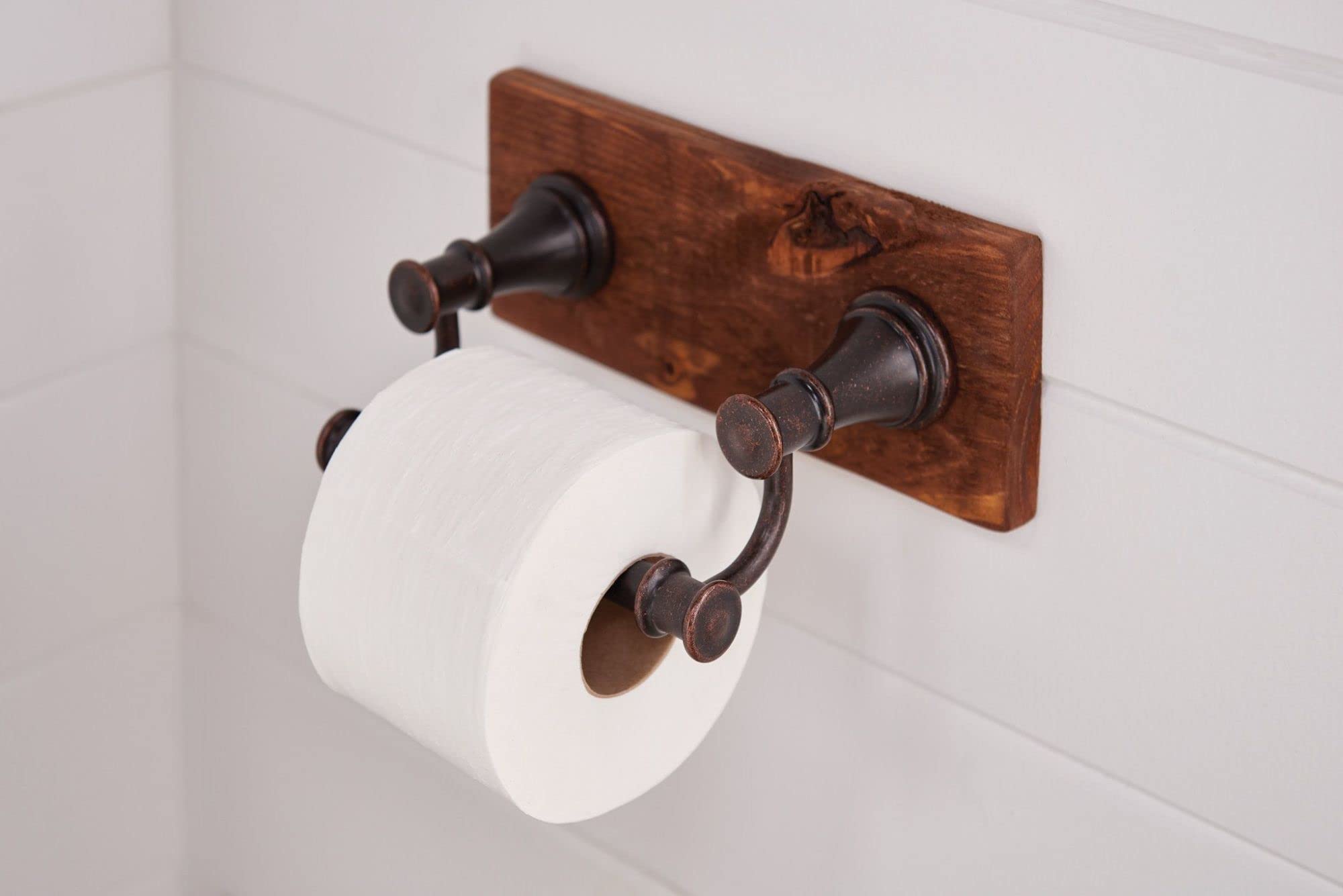 Oil-rubbed bronze toilet paper holder with curved arm mounted on wooden backing plate holding white roll.