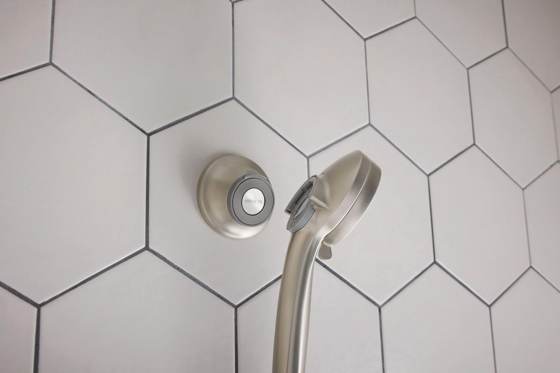 Brushed nickel showerhead with magnetic remote cradle mounted on hexagonal tile wall.