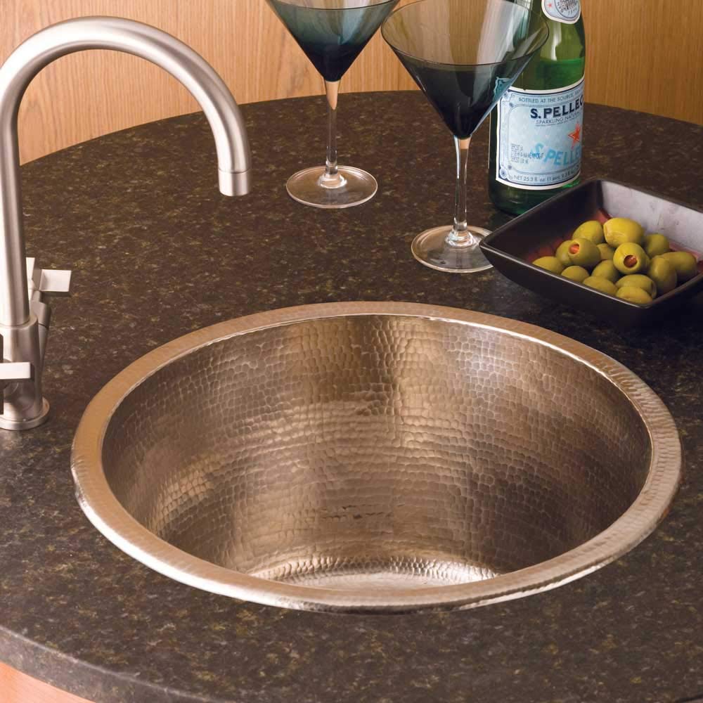 Hammered copper round bar sink with curved faucet installed in dark countertop with cocktails and garnishes nearby.