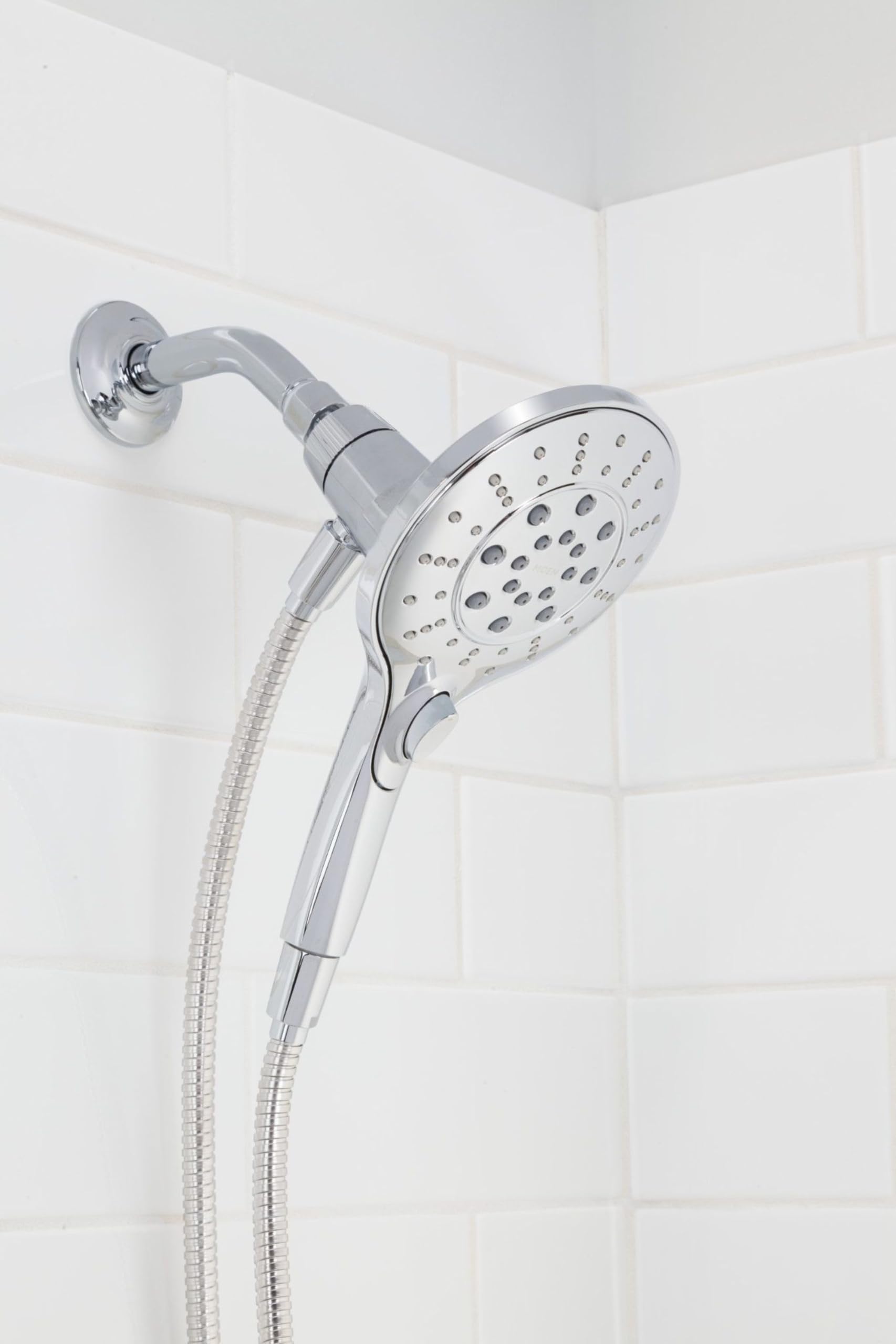 Chrome handheld showerhead with round spray face and flexible hose attached to wall mount on white tile bathroom wall.
