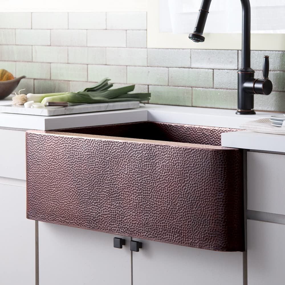 Antique copper farmhouse sink with hammered front apron installed under white countertop with black faucet.